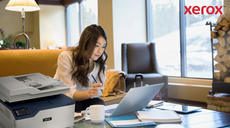 Piattaforma Xerox® Workflow Central: lavora ovunque Una donna lavora al computer portatile in un comodo ufficio o in una sala relax, con una stampante compatta Xerox visibile in primo piano, a indicare la perfetta integrazione della stampa con i flussi di lavoro digitali.