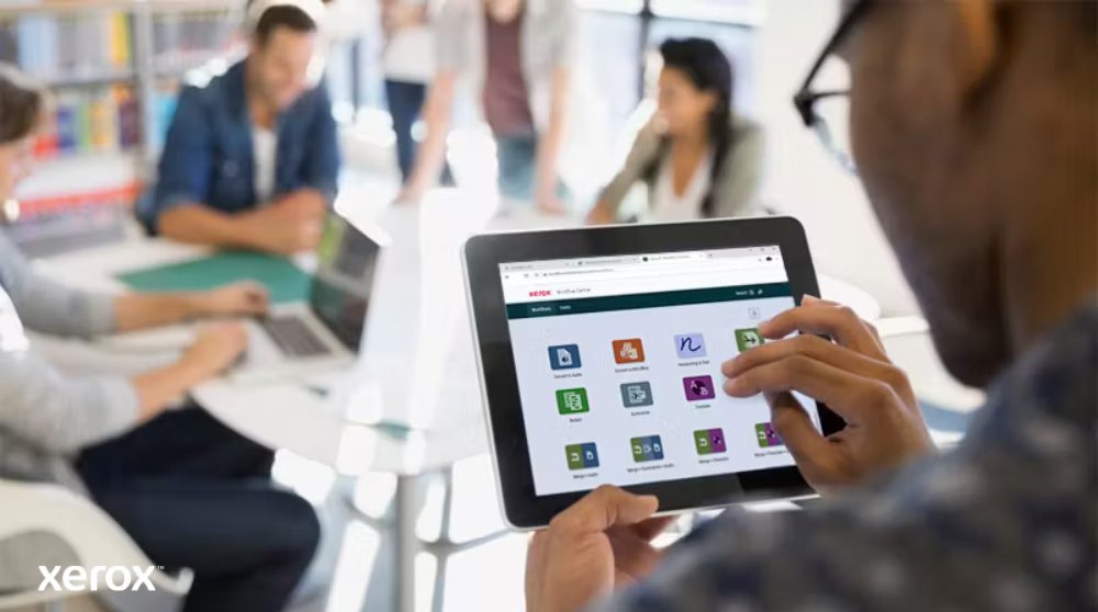 Piattaforma Xerox® Workflow Central: accesso mobile intuitivo Una persona tiene in mano un tablet che mostra l'interfaccia di Xerox Workflow Central con varie icone di app, mentre altre persone lavorano sullo sfondo sfocato, evidenziando l'accesso mobile alle soluzioni di flusso di lavoro.