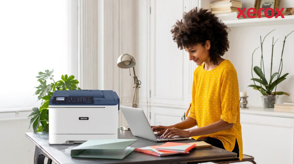 Una persona con i capelli ricci sorride mentre scrive su un computer portatile seduto alla scrivania, con una stampante compatta Xerox C310 posizionata accanto al computer portatile, a dimostrazione di una moderna configurazione di ufficio domestico per la produttività.