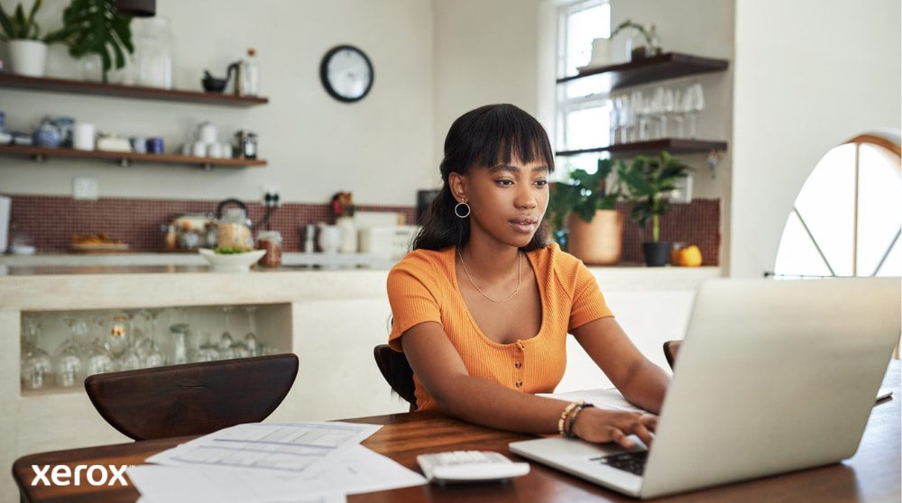 Una persona è seduta al tavolo della cucina, concentrata sul lavoro con un computer portatile, con documenti e una calcolatrice a portata di mano, a dimostrazione della flessibilità del lavoro da casa con una gestione efficiente dei documenti.