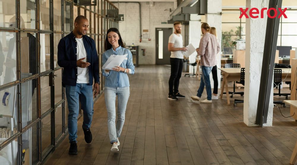 Xerox® Print and Scan Experience: potenziare la collaborazione sul posto di lavoro Due colleghi camminano lungo un corridoio in un moderno ufficio open space, tenendo in mano dei documenti e sorridendo, con altri colleghi visibili sullo sfondo, a rappresentare un ambiente di lavoro dinamico e collaborativo.