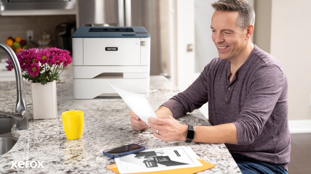 "Esperienza di stampa e scansione Xerox®: gestione dei documenti senza sforzo " Una persona sorride mentre esamina un documento seduto al bancone della cucina, con una stampante compatta Xerox e una tazza gialla brillante nelle vicinanze, a suggerire una stampa e una gestione dei documenti senza problemi in un ambiente domestico o in un piccolo ufficio.