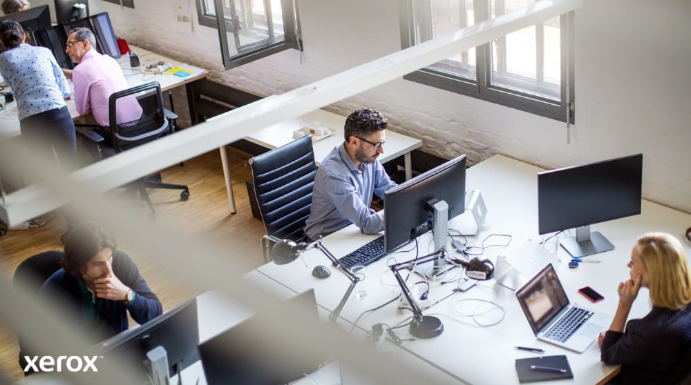 Xerox® Print and Scan Experience: aumentare la produttività in ufficio Una vista dall'alto di un ufficio open space con diverse persone che lavorano alle loro scrivanie, utilizzando i computer. La scena trasmette un ambiente di lavoro produttivo e collaborativo.
