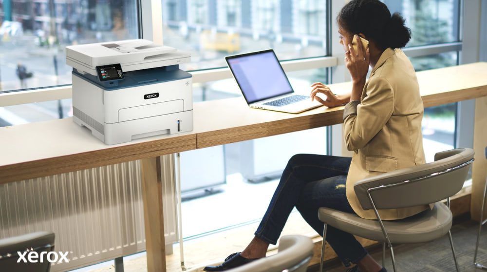 Esperienza di stampa e scansione Xerox®: comoda e connessa Una persona è seduta a un tavolo alto vicino a una grande finestra, mentre parla al telefono e lavora al computer portatile. Una stampante multifunzione Xerox B235DNI è comodamente posizionata sul tavolo accanto, a suggerire la facilità d'uso in un ambiente di lavoro moderno.