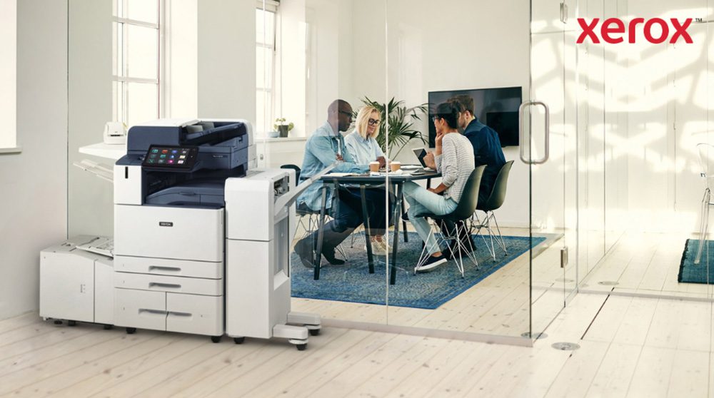 Due persone sedute a una scrivania, con le spalle rivolte alla telecamera, si salutano con un pugno, simbolo di lavoro di squadra e collaborazione di successo. Una di loro guarda lo schermo di un computer. Una moderna stampante multifunzione Xerox AltaLink B8245 è in primo piano in un ufficio luminoso. Sullo sfondo, un gruppo di persone è impegnato in una discussione collaborativa attorno a un tavolo.