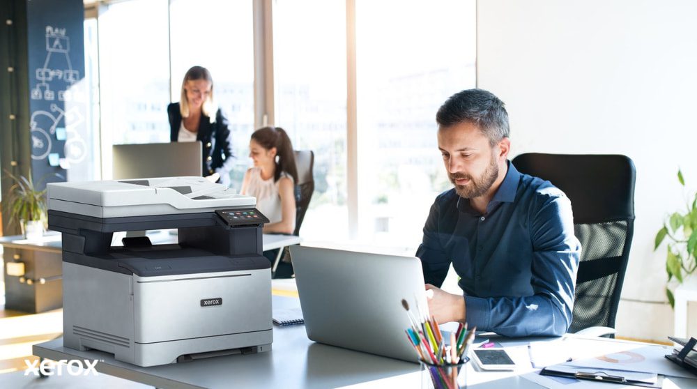 Xerox® Smart Start: progettato per gli spazi di lavoro moderni Una persona è seduta a una scrivania e guarda un computer portatile con accanto una stampante multifunzione Xerox C325. Sullo sfondo, altre due persone lavorano alle loro scrivanie in un ambiente luminoso.