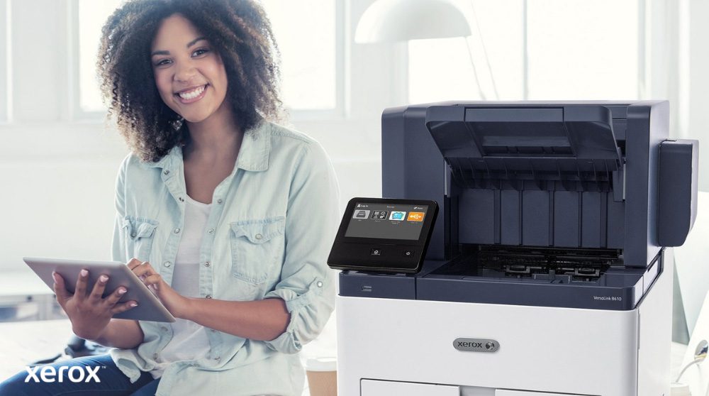 Xerox® Workplace Cloud e Suite: integrazione intuitiva Una persona sorridente è seduta accanto a una stampante bianca Xerox VersaLink B610, tiene in mano un tablet e guarda la telecamera, evidenziando la natura intuitiva delle soluzioni Xerox.