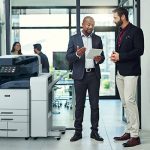 Due uomini in un ufficio discutono di qualcosa su un tablet accanto a una stampante multifunzione a colori Xerox® AltaLink® serie C8200 Due uomini in un ufficio discutono di qualcosa su un tablet accanto a una stampante multifunzione a colori Xerox® AltaLink® serie C8200