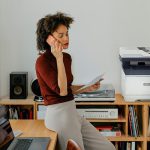Stampante multifunzione a colori Xerox® C325 in un ambiente di ufficio domestico Una donna seduta alla scrivania mentre fa una chiamata accanto a una stampante Xerox® C325