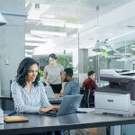 Stampante multifunzione a colori Xerox® C325 in una situazione d'ufficio Donna professionista che digita sul laptop alla scrivania in ufficio accanto a una stampante Xerox® C325