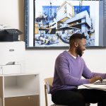 Stampante multifunzione Xerox® VersaLink® C415 sul posto di lavoro Uomini che lavorano su un computer portatile davanti a una stampante multifunzione a colori Xerox® VersaLink® C415