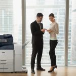 Due persone in un ufficio in abiti da lavoro che discutono di documenti sono in piedi accanto a una stampante multifunzione Xerox® AltaLink® serie B8200 Due persone in un ufficio in abiti da lavoro che discutono di documenti sono in piedi accanto a una stampante multifunzione Xerox® AltaLink® serie B8200