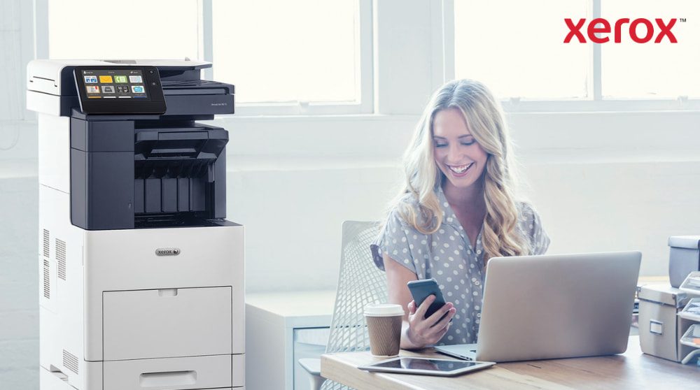 Xerox® ConnectKey®: integrazione perfetta del flusso di lavoro Una persona guarda felicemente il suo smartphone mentre è seduta alla scrivania con un computer portatile. Una stampante multifunzione Xerox VersaLink B605 è visibile accanto alla scrivania, in un ufficio ben illuminato.